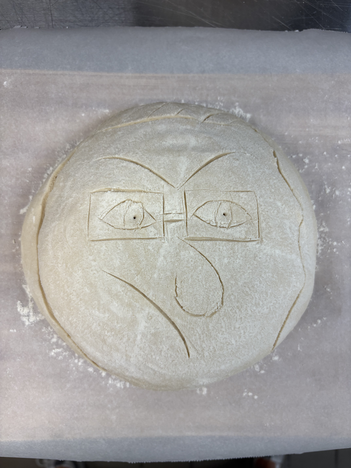 Sourdough loaf scored with a bespectacled face shedding a single tear, cooling on a wire rack — alternate view