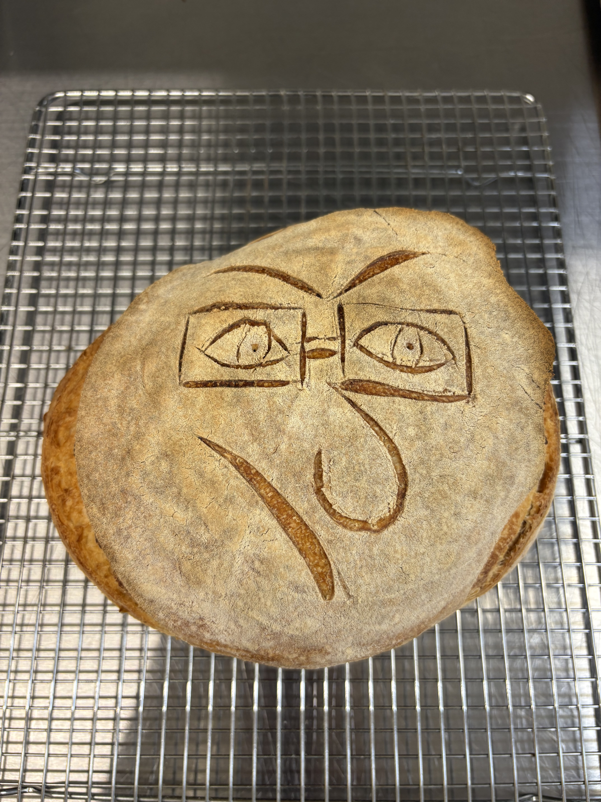 Sourdough loaf scored with a bespectacled face shedding a single tear, cooling on a wire rack