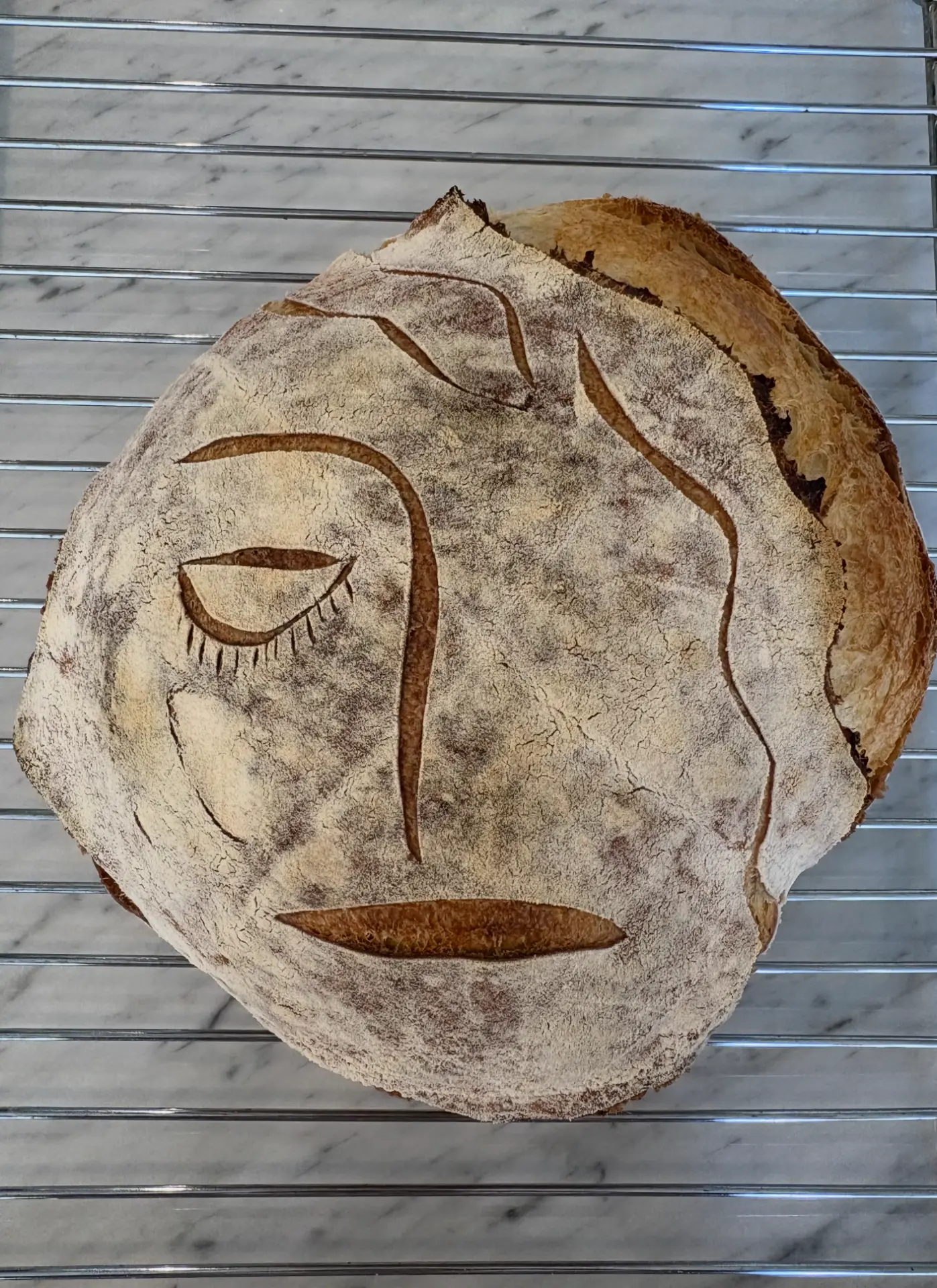 Sourdough loaf scored with a serene closed-eye face, styled with rosemary and olives — alternate view